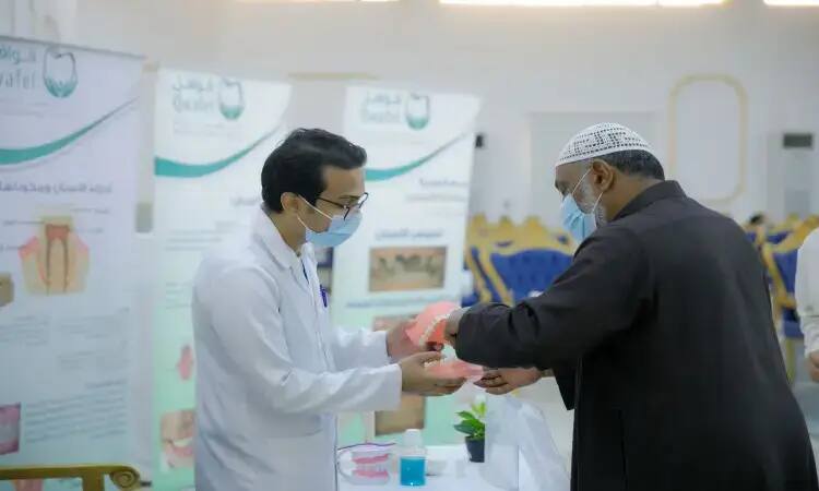 A doctor in a white coat advises dental hygiene using a Yee model in front of a man wearing a Saudi shemagh with an aqal and bisht, at an informational booth organized by the Qassim Dental Caravans, with awareness posters in the background.