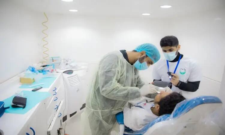 A dentist dressed in a Saudi bisht and ghutra is examining a child's teeth in a dental center clinic, while an assistant from the Qassim Dental Caravans, wearing an aqal and a Saudi ghutra, is at his side taking notes. The clinic appears clean and well-lit, with medical equipment on display.