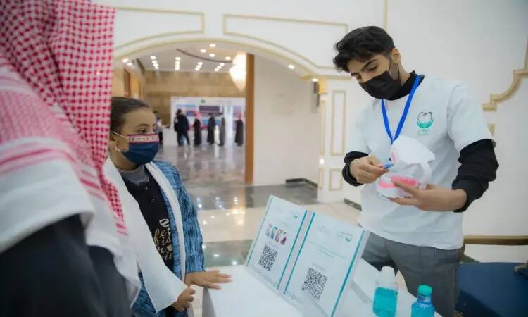 One of the organizers wore a Saudi-style ghutra, a black mask and a white shirt. This person is demonstrating dental care methods using a large model of a tooth in front of children during a health education event held at Qubba Center and organized by Qassim Dental Caravans. A table of awareness materials and a number of health education vials are placed on the table.