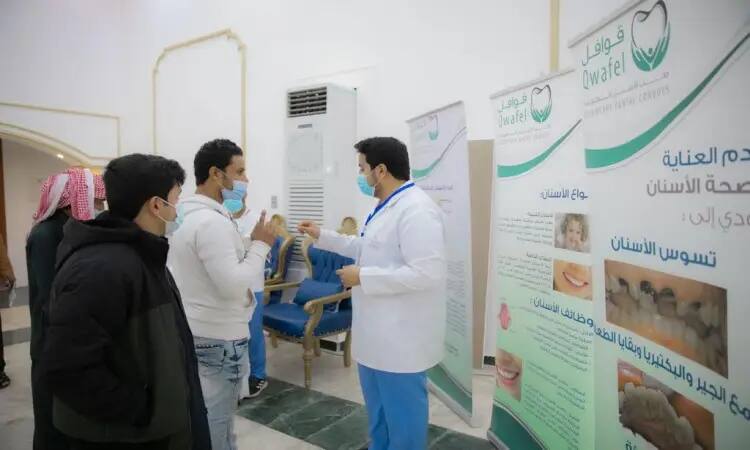 A dentist in a white coat and muzzle chats with three men in the Saudi Arabian ghutra or shemagh, aqal and bisht, thanks to a sign informing them about oral and dental hygiene, at an event held at the Qassim University Dental Caravan Center. During the conversation, one attendee gestures with his hand while the rest pay attention; a sticker displays tips in Arabic for a clear bottle toothbrush.