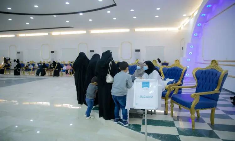 A Florida group of women wearing black abayas, accompanied by several children, at the humanitarian aid desk in a brightly lit hall with blue chairs at the University Dome Center in Qassim. A small section of a sign with text in Arabic introduces the dental caravans initiative. Some of the men were dressed in the Saudi shemagh or white ghitra with the Saudi aqal and bisht, signaling the authenticity and national heritage of the place.