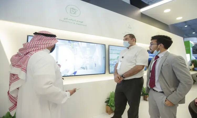At the "Prevention and Treatment" pavilion during the Middle East Poultry Exhibition in Riyadh, three men exchange pleasantries; two of them are dressed in long-sleeved clothing, while a third is wearing a Saudi tunic and men's shirt with an official university certificate including a national ID. Two of the attendees are wearing masks while parked in front of a large projection screen at the booth.