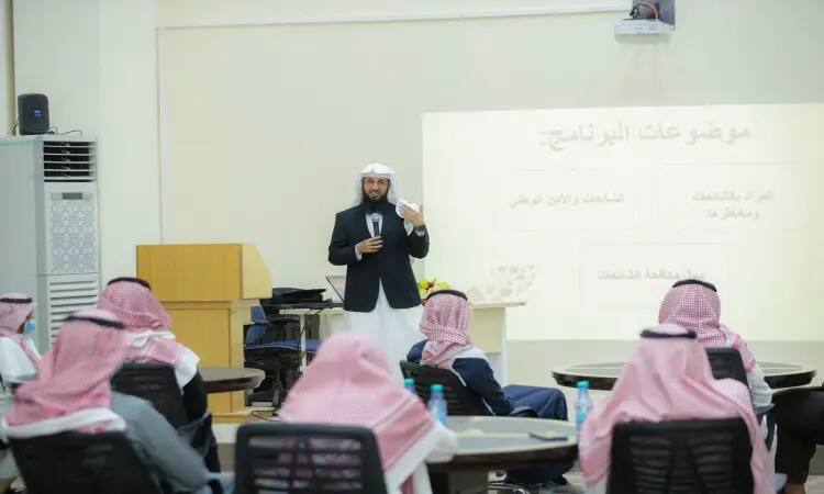 يقدم رجل يرتدي الزي السعودي الرسمي، المكون من غترة أو شماغ وعقال بالإضافة إلى بشت سعودي، عرضًا حول موضوع التحصين الفكري أمام مجموعة من الرجال الذين يستحقون الغترة أو الشماغ والعقال ويجلسون حول طاولات مستديرة داخل القاعة. السيطرة على المحاضر على الشاشة نص باللغة العربية لعلاج موضوع "تشخيص".