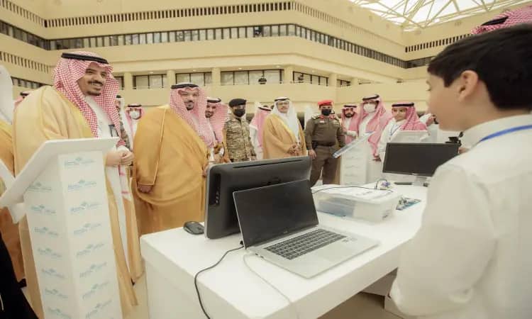 الاتصال مجموعة من الرجال مرتدين الغترة أو الشماغ والعقال والبشت السعودي، يرافقهم أعضاء الأمن، حول منصة لمس بأجهزة حاسب آلي، وهم يتفاعلون مع شاب ضمن فعاليات معرض مبتكر 2022 الذي نظمته جامعة القصيم في إحدى القاعات الداخلية.
