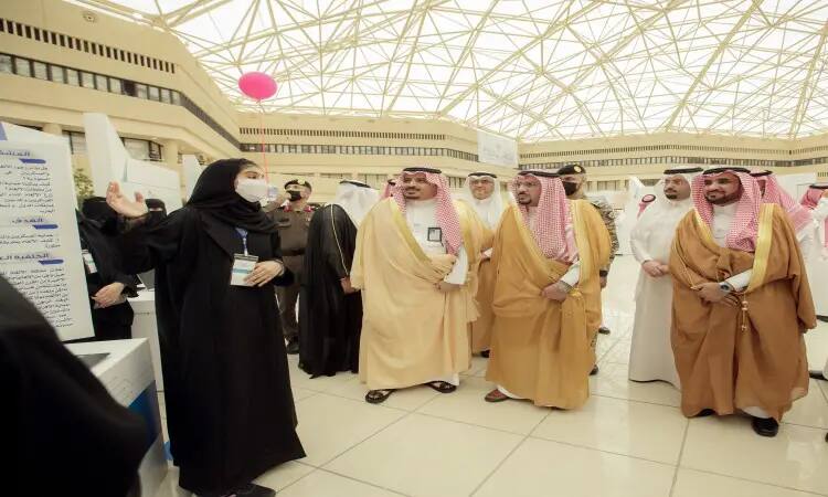 يجتمع عدد من الرجال مرتدين الغترة أو الشماغ والعقال، إضافة إلى البشت السعودي، للاستماع إلى سيدة ترتدي ملابس بيضاء وهي متقدمة عرضاً تقديمياً ضمن فعاليات معرض داخلي ذي زجاجي هندسي، وذلك من خلال فعالية "مبتكر 2022" في جامعة القصيم.