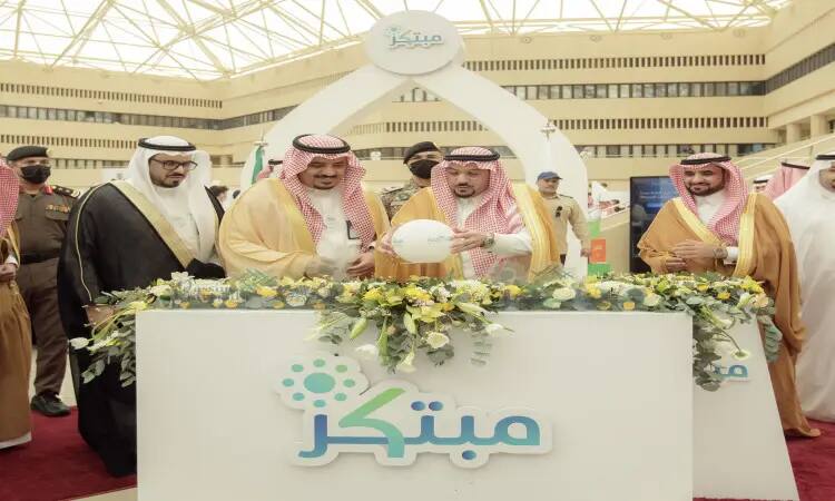 الاتصال عدد من الرجال مرتدين الغترة أو الشماغ السعودي مع العقال، بالإضافة إلى البشت السعودي التقليدي، حول منصة عرض تحمل باقات من الزهور وشعار "مبتكر" ضمن فعاليات معرض مبتكر 2022 في جامعة القصيم. نورت في الخلفية حضور منسق تحت السقف الزجاجي العالي، بما في ذلك التوقيع الرسمي والمميز للعرض.