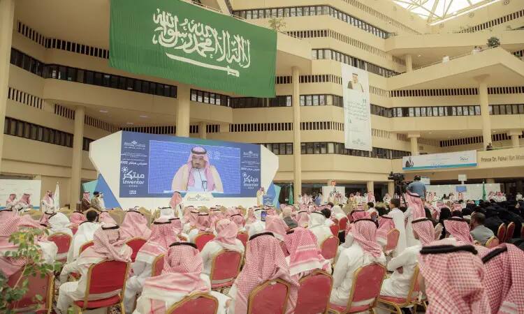 يشارك جمع كبير من الرجال مرتدين الغترة أو الشماغ السعودي مع العقال والبشت السعودي في مؤتمر رسمي يُعقد داخل جامعة القصيم. أخيرًا على المسرح، أصبح كبير الحجم أن يكون أمير منطقة القصيم، يلقي كلمته بحضور العلم السعودي وشاشة كبيرة خلفه تم عرض صورته ضمن هذا الحفل الرسمي.