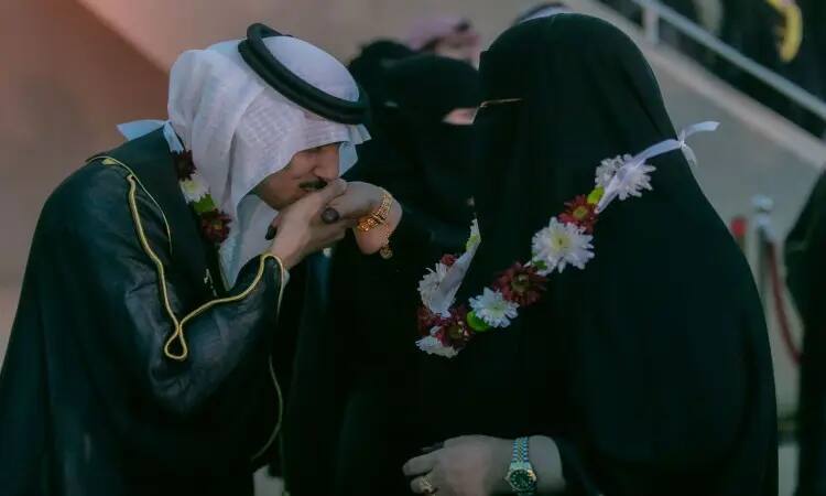 خلال حفل تخرج الفوج التاسع عشر بجامعة القصيم، شوهد رجل يرتدي البشت السعودي التقليدي والغترة والعقال وهو يُقبّل يد امرأة ترتدي عباءة سوداء ونقابًا، وكلاهما مُزيّن بأكاليل الزهور. تظهر شخصيات مُشوّشة في الخلفية، مُسلّطةً الضوء على هذه اللحظة المؤثرة التي تشاركتها الأمهات في هذه المناسبة المُميّزة.