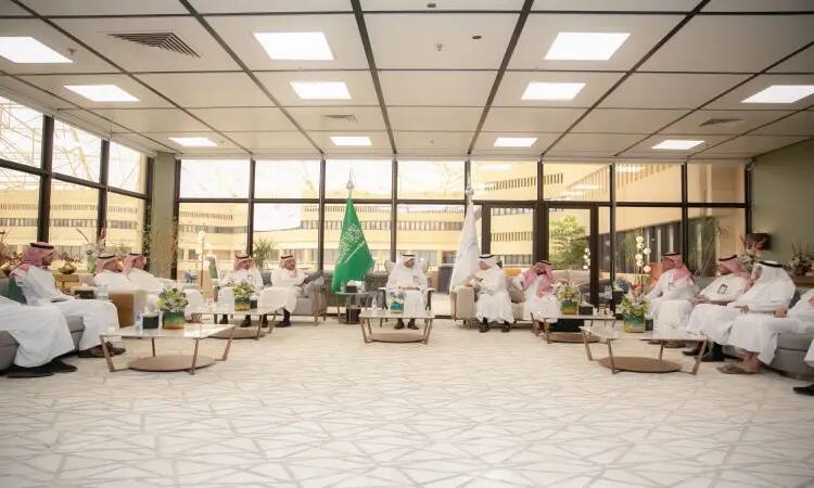 A number of men dressed in the Ghutra or Shemagh and Eqbal, some in the official Saudi winter dress, are gathered in a large modern hall with large windows. Saudi Arabia's flags adorn the hall and flowers are placed on the tables as they hold key discussions as part of the Qassim government workshop on energy conservation.