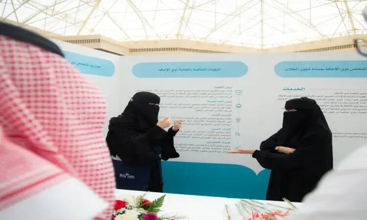 Two women are communicating outside the information booth of the Deanship of Student Affairs at Qassim University, where the Ruwwad program is featured. The man next to them is wearing a Saudi bisht, shemagh and aqqal. Eugene is adorned with Arabic phrases and flowers, in support of the Ruwwad program.