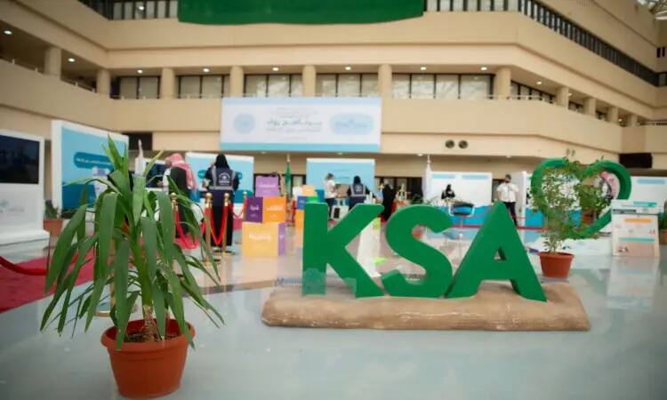 Huge sculptures with prominent green letters of the word "KSA" are centered in an indoor exhibition hall, prominently displayed in pots. Notice the prominent presence of people wearing the Saudi Ghutra or Shemagh, Eqbal, and Bisht, including confirmation of national identity. Surrounding the exhibit are several informational booths for the sponsors and the Deanship of Student Affairs, indicating the organization of the Qassim University event to support the winners by contributing to university assistance while respecting Saudi traditions and values.