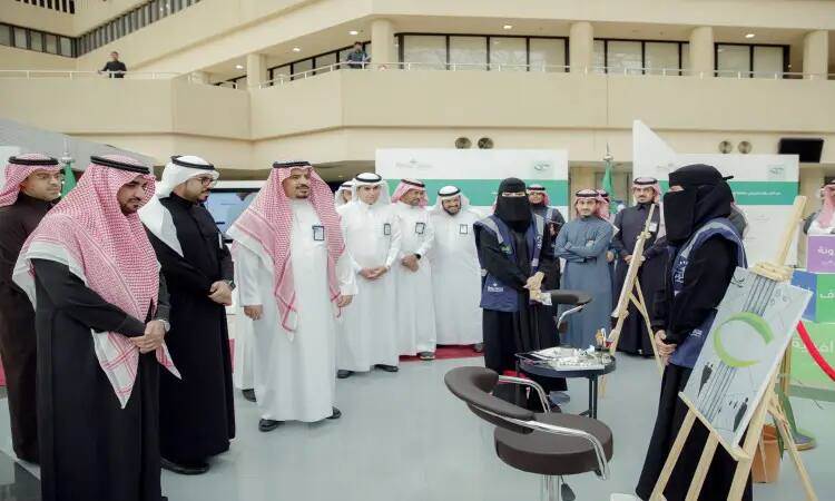 تجمّع مجموعة من الرجال قمصان الشماغ والعقال والبشتاء السعودي، وعدد من النساء داخل عباية السيدات قاعة مغلقة، حيث تتابع الحضور باهتمام عرضًا فنيًا تبرزه ملابس أمام منصة للرسم. وتبرز في الخلفية أجنحة الكون التي تسلط الضوء على جهود المملكة وبرنامج نزاهة في مكافحة الإرهاب.