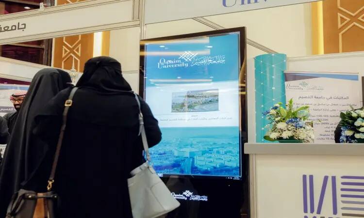 Florida, USATwo women in black hijabs stand next to a large digital display screen at the Qassim University pavilion during the Qassim Book Fair. The screen disseminates the university's information and publications, while brochures and flower arrangements are scattered around the area. One of the university's licensees, dressed in a Saudi Ghutra with a Saudi Eqal and Bisht, was licensed in September to reflect the official character and national identity of the Qassim University pavilion at the trials.