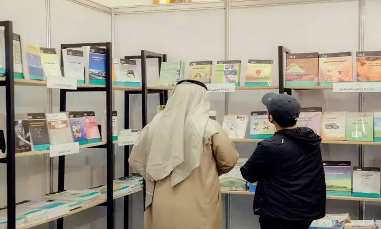 In front of black metal shelves adorned with the university's publications in the center of the Qassim Book Fair. Arabic books and magazines are stacked on shelves lined with a white wall, reflecting the scientific and cognitive excellence of Al-Qassim University.