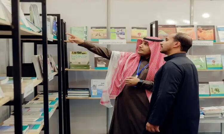 An Altans man in front of shelves laden with books at the Qassim Book Fair; a Saudi jacket, headband and bisht must have helped him to one of the books implicitly, while the other man got the same mistake.