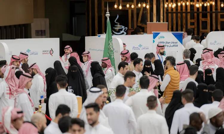 A large gathering of men and women gather in a closed hall during a technical career practice organized by Qassim University, where many attendees bring the Saudi Ghutra or Shemagh with Eqbal, and some participants share the traditional Saudi Bisht. To complete the pavilions, distinctive banners and the Saudi Arabian flag were displayed. Attendees engaged in discussion and communication with representatives of different organizations, as their conversations related to topics related to technical information and job opportunities in this field.