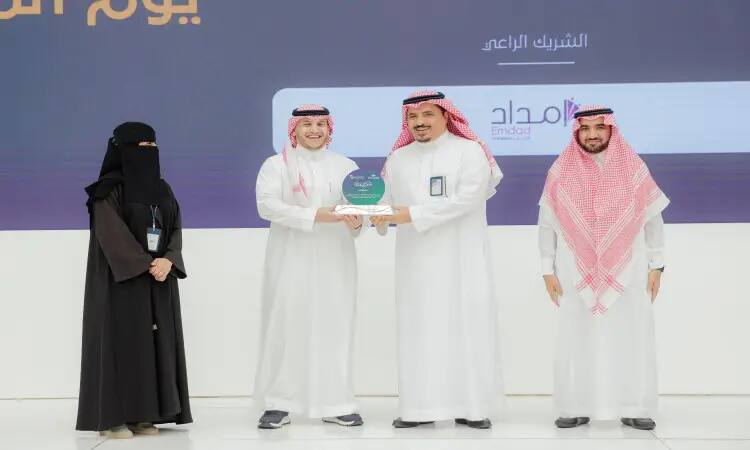 During a professional art audition, four people on stage at an award ceremony, one of the men handing an award to another man. A woman dressed in women's clothing and a black niqab stands on the left, while two men dressed in Saudi robes with shemagh, headband and Saudi bisht, representing the Ministry of Communications, are standing next to her.