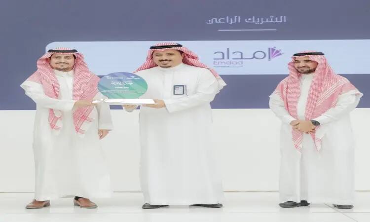 Three men call to the stage dressed in white, two of them wearing the Saudi shemagh and headband while the third wears a Saudi bisht over his dress with the shemagh. Two of the attendees are handing out a joint honorary annual shield, while the third man next to them is dignified. A reminder of Qassim University logos, a logo of supplies and phrases in Arabic referring to "Technical Profession Day."