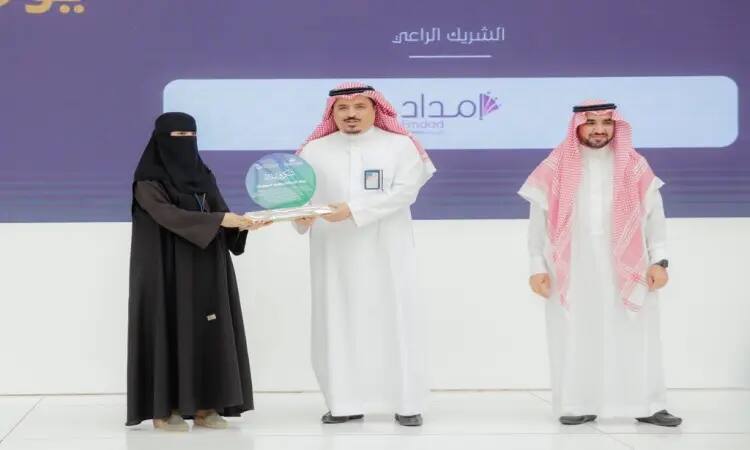 A man wearing a white suit and a Saudi besht over the white outfit, with a Saudi shemagh and a Saudi ghutra, received the award during the Technical Career Day activities at Qassim University. A representative from the Ministry of Telecommunications, also dressed in a white suit and a Saudi besht with a Saudi ghutra, stood beside them, smiling during the restoration.