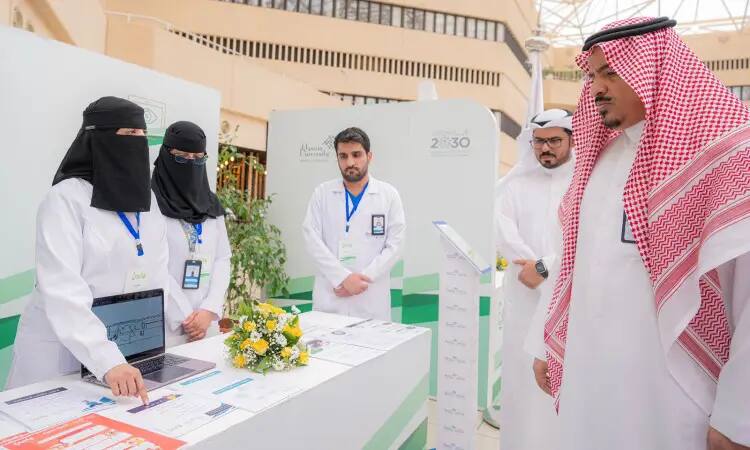 Contact a group of healthcare professionals, handing out white coats, at an introductory corner at the Wai Exhibition in Qassim. A man dressed in a Saudi Arabian ghutra, aqal and bisht listens to a briefing on a computer and documents.
