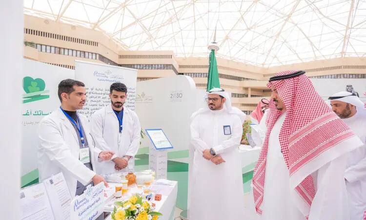 A group of men wearing the Saudi Ghutra, Shemagh, Egal and Bisht, along with a number of medical staff members in white coats next to a table containing awareness products about drug detection, in addition to flowers and some medical supplies, within the activities of the "Wai2" exhibition at Qassim University, and following in the background the Saudi flag and a number of introductory kits for the exhibition.