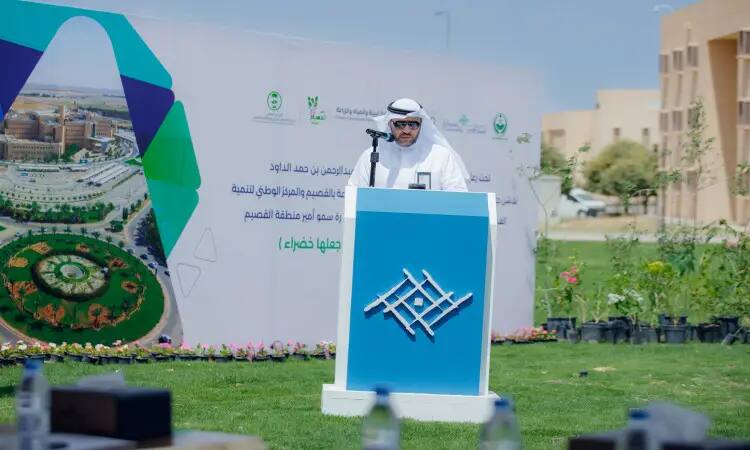 رجل يرتدي عادة الغترة السعودية، العقال، البشت السعودي والشماع، أمام منصة في الهواء الطلق خلال فعالية نظمتها جامعة القصيم. ويظهر خلفه شعار مبادرة "لنجعلها خضراء" مع المساحات الخضراء، مما يشير إلى إمكانية العمل على جهود التنمية الحضرية والمحافظة على البيئة.