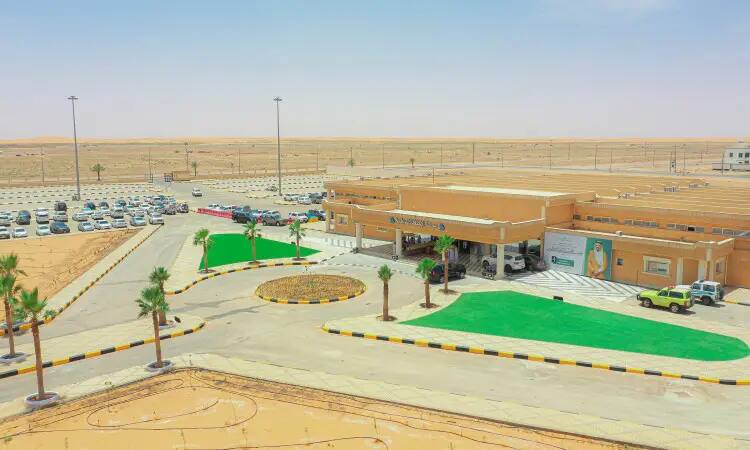 A new hospital building with parking lots, green spaces of artificial grass, palm trees and cars, a branch in the heart of the desert under a clear blue sky. This image shows a young man wearing a Saudi official Ghutrah, Shamma'a, and Bisht, symbolizing his authenticity and reflecting the national identity. This visual represents the Olive Health and Environmental Awareness and Education Campaign organized by Qassim University, highlighting the importance of a healthy environment and authentic Saudi identity.