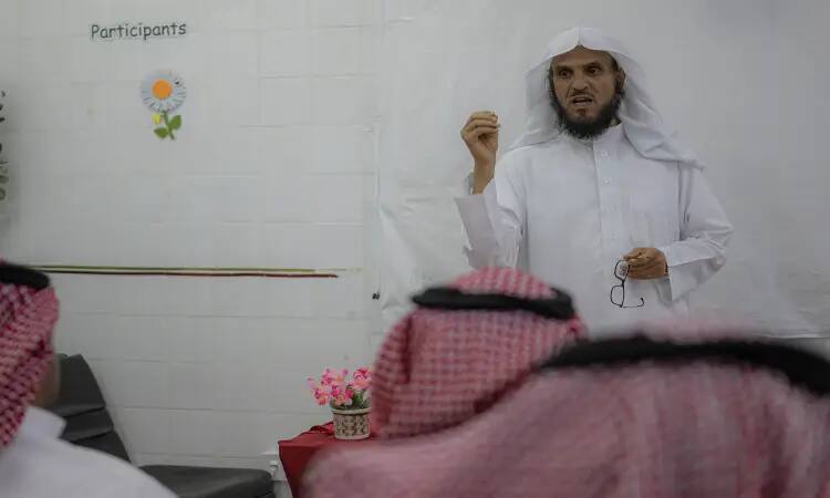 A cat man dressed in a Saudi Ghutra with aqal and Saudi Bisht in front of the audience, participating in the health, awareness and education campaign at Al-Qassim University. He squeezes his glasses as a number of people listen to him, the Saudi shemagh lagging behind him, a whiteboard with the word "Participants" written on it.