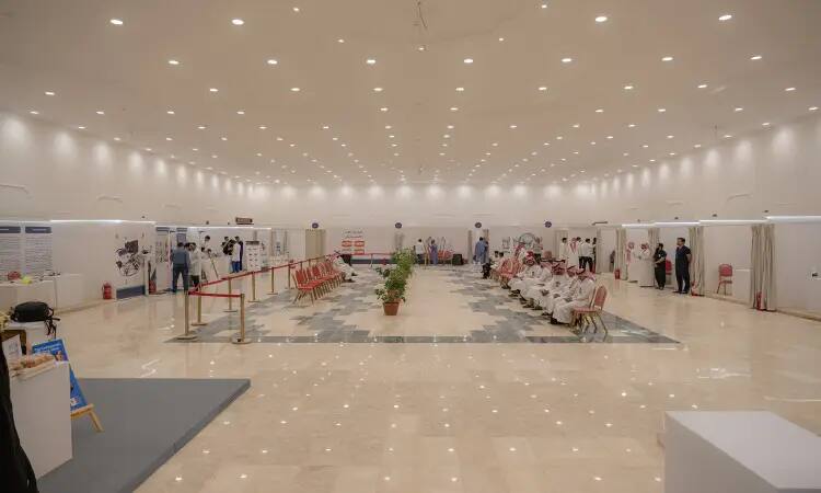 A brightly lit hall, with attendees seated on one side of it, the group is represented by the Saudi Ghutra or Shemagh, Eqal, and Bisht, including the official national attire. There are several specialized sections of the outreach pillow at the edges of the pavilion, where it is located in a decorative base that adds an aesthetic touch. The team provides support to the participants from the staff and students of Qassim University.