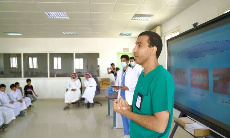 A man in a green medical uniform delivers oral health awareness information via a projection screen to a number of students and a number of parents seated in the auditorium. The scene is underscored by members of the military warriors in white uniforms and masks, as well as a number of attendees in formal Saudi attire, wearing the katira or shemagh and aqal, and some in the Saudi bisht, including a full confirmation that it is Qassim University.