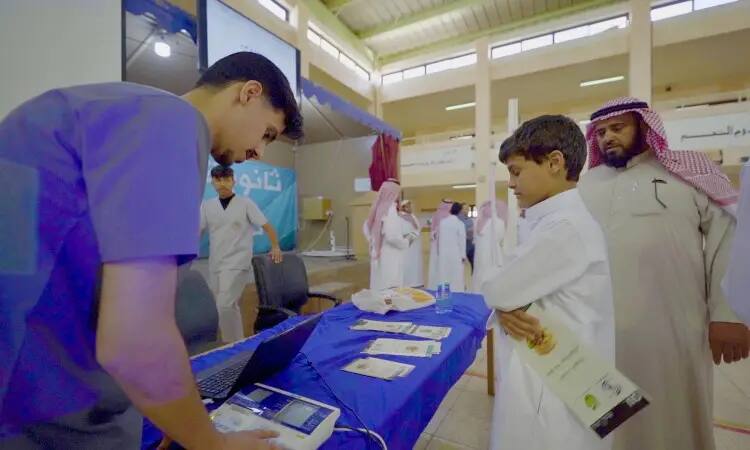 A health care worker dressed in a medical uniform provides health education to a young child at a medical center based in a public place. A number of men are wearing the Saudi thobe and shemagh or the Saudi ghutra with an aqal, and some figures are wearing the Saudi bisht, and health awareness pamphlets are displayed in an organized manner during Blue Week.