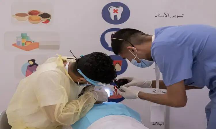 Two medical specialists, wearing masks and necessary supplies in addition to the Saudi official Ghutra, Eqal and Bisht, examine a patient using a hand-held light inside a clinic, as part of the activities of the Seha campaign, which aims to raise health awareness. The poster on the back of the clinic features the logo of Al-Qassim University.
