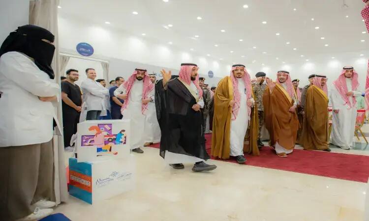 The Emir of Qassim, dressed in a Saudi bisht, walks the red carpet among attendees in a brightly lit hall. Inside the venue, the woman is not wearing a white coat and is covered by a Saudi bisht near a colorful display case, as part of the awareness and education programs organized by Al-Qassim University.