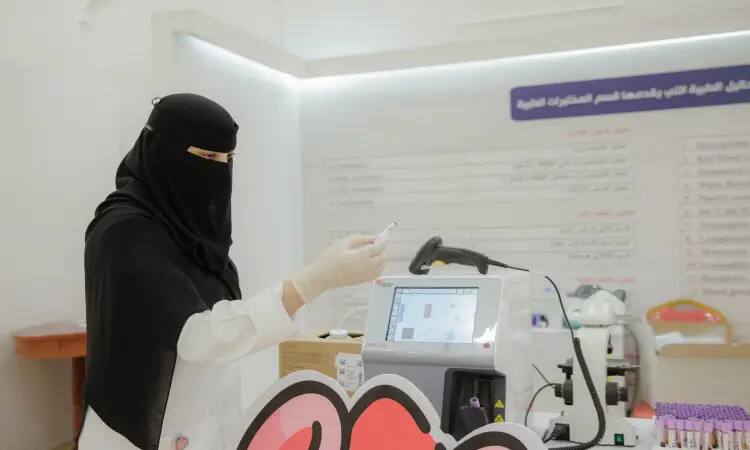 ملابس رياضية سعودية ترتدي نقابًا بلابازات، كما ترتدي ملابس وفوقها عقال، بالإضافة إلى بشت سعودي رسمية، تتحمل اختبار الأنبوب أثناء العمل مع أجهزة المختبر في بيئة سرير داخل جامعة القصيم، لتعزيز التوعية الصحية. ضع خلفها على الحائط مخطط يحتوي على نصوص عربية.