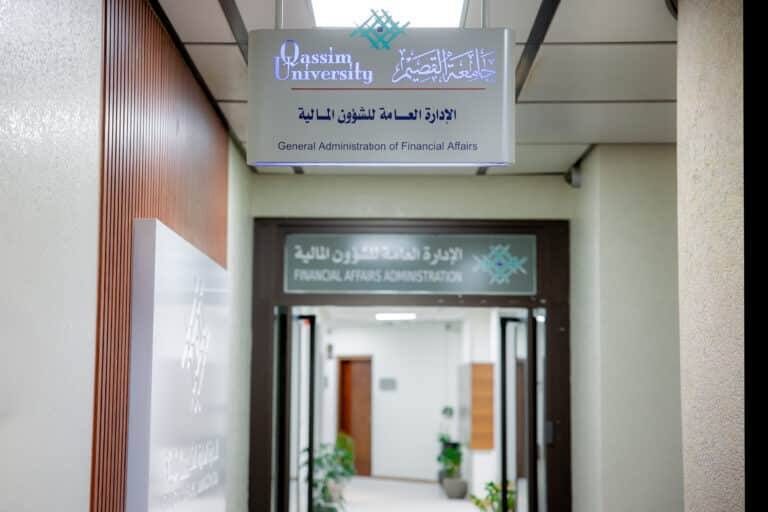 توجد في أحد أروقة جامعة القصيم لافتة علوية مكتوب عليها "الإدارة العامة للشؤون المالية" باللغتين العربية والإنجليزية، ترشد الزوار إلى مكتب الإدارة المالية. يتواجد أفراد يرتدون الزي السعودي التقليدي مثل الثوب مع الشماغ أو الغترة. تعكس الأجواء بيئة جامعية رسمية ومهنية.