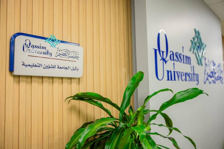 نبتة موضوعة موضوعة أمام جدار يحمل شعار جامعة القصيم ونمطًا باللغة العربية بالإضافة إلى لوحة تشير إلى مكتب الشؤون التعليمية التابع للجامعة.