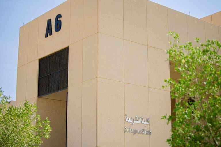 مبنى ذو لون بيج يحمل الرمز أ6 واسم كلية الشريعة باللغتين الإنجليزية والعربية، ويحيط به أشجار خضراء تحت سماء صافية زرقاء.