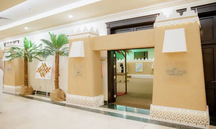 The entrance to a distinctive building of traditional Arabic architecture, highlighted by its shiny walls and geometric patterns, is surrounded by heritage-era industrial palm trees. Two glass banners with Arabic text adorn the entrance, near a door to the university's exhibition at the Buraydah Dates Carnival. In front of the building, a number of visitors appear wearing the Saudi shemagh, aqal, besht or ghutra, signaling the region's official identity and reinforcing the heritage values that Qassim University is proud of.