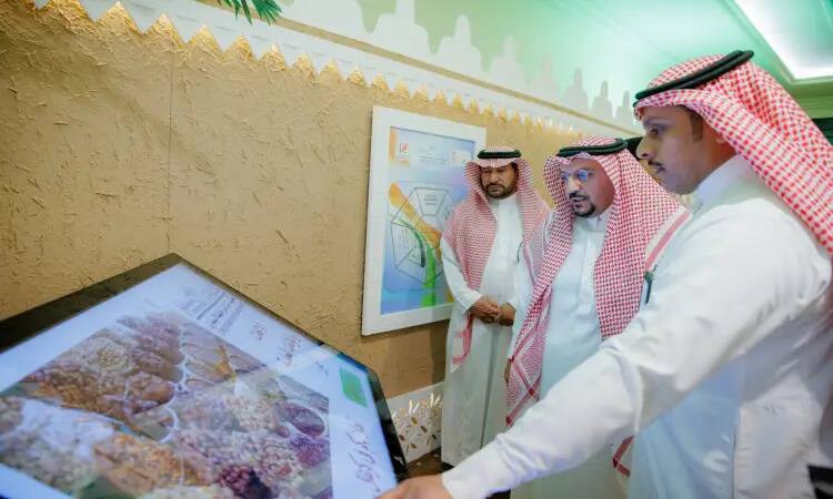 Three men in Saudi Pashtuns, Ghutra or Shemagh and Egal using a large interactive screen where various types of grains were spotted, as part of the Buraydah Dates Carnival. In the background, awareness posters about the date palm and infographics were displayed on the wall, including an educational and organizational printer provided by Qassim University.