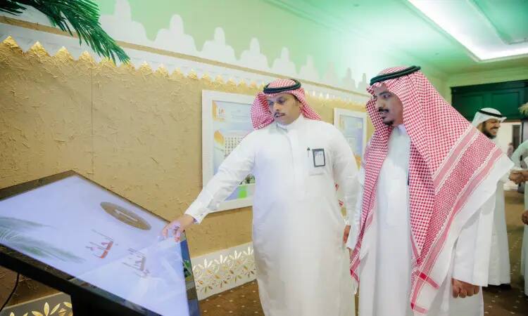 يتفاعل رجلان يرتديان الزي السعودي الرسمي الامتداد في الغترة أو الشماغ والعقال بالإضافة إلى البشت السعودي مع شاشة تفاعلية كبيرة في جناح جامعة القصيم خلال كرنفال بريدة للتمور، وجزء في الخلفية عدد من الحضور وبعض العناصر الزخرفية.