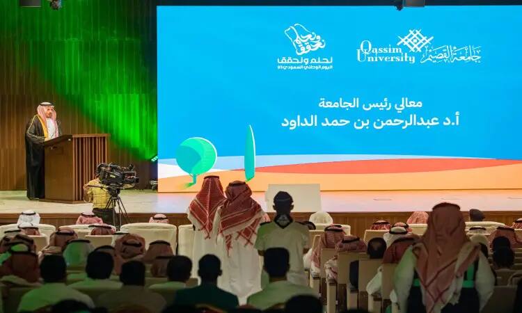 He takes the podium in front of the audience at the event, with a large screen behind him bearing the Qassim University logo and a text in Arabic about National Day. In the hall, a number of attendees are wearing shemagh or ghutra and headband, in addition to the Saudi bisht, as a sign of national identity and the official traditions of the occasion.
