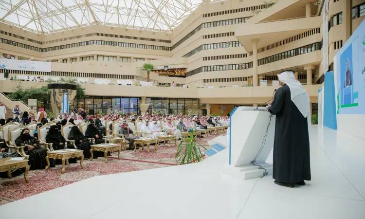 رجل يرتدي الغترة السعودية والعقال، وقد اكتسى بالبشت السعودي الرسمي، تومكاتا من على المنصة خلال فعاليات الملتقى للجامعة. يخاطب في كلمته جمعًا متنوعًا من الحضور، من بينهم أعضاء أندية نزاهة السفر، وذلك في ردهة خارجية والآن بتصاميم معمارية حديثة ونوافذ واسعة في القصيم.