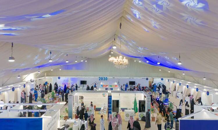A wide view of an indoor exhibition held at the Mudkhana Center, the exhibition is organized under a large tent decorated by Lart. It contains multiple elements in corners dealing with awareness, painting and education, and includes a large number of visitors who noticed the Saudi tunic, shemagh, headband and bisht, with a clear logo of Vision 2030 in the background.
