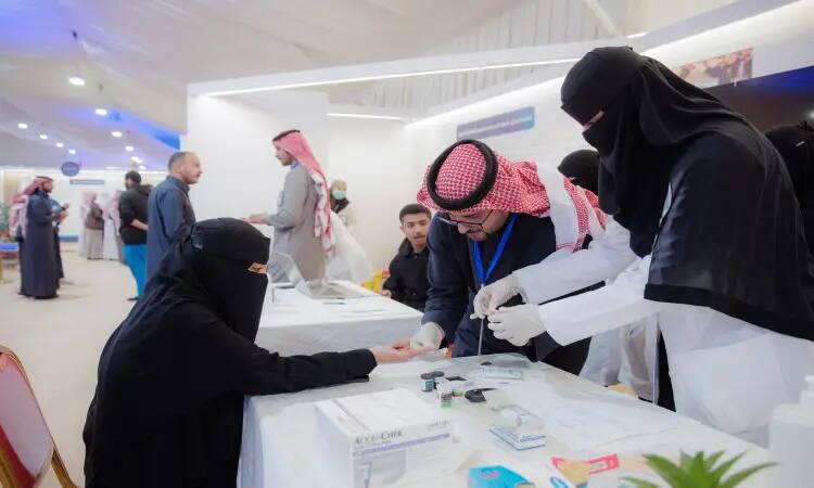 Dressed in white and niqabi clothes, she sits on a table in a medical ward at Dukhna Center, where a health protection worker is testing her blood as part of awareness and education activities. In the background are a number of attendees wearing the Saudi bisht, Saudi shemagh or Saudi ghutra with an aqal, in a picture of respect for formality and adherence to the authenticity of the national dress code inside Qassim University.