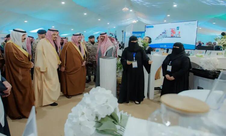 A group of men dressed in Saudi Arabian ghutra or shemagh with aqal and besht, along with two women dressed in white and niqab, connect inside a tent set up for a health awareness presentation at Qassim University. She gestures as she speaks, while projecting a large screen at the site.