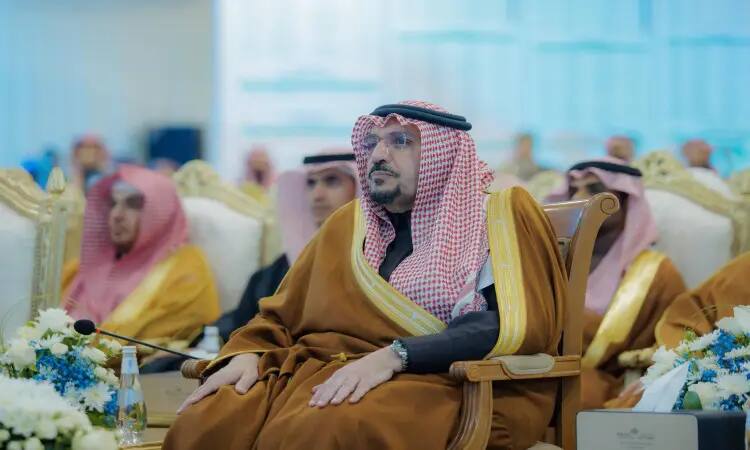 A man stands, wearing a Saudi Arabian khalqal and bisht over his shamma, representing the Emirate of Qassim, on a plush chair during a cultural and educational outreach event. In front of her are the coordinating formats and water bottles at the official ceremony held at the University of Qassim.