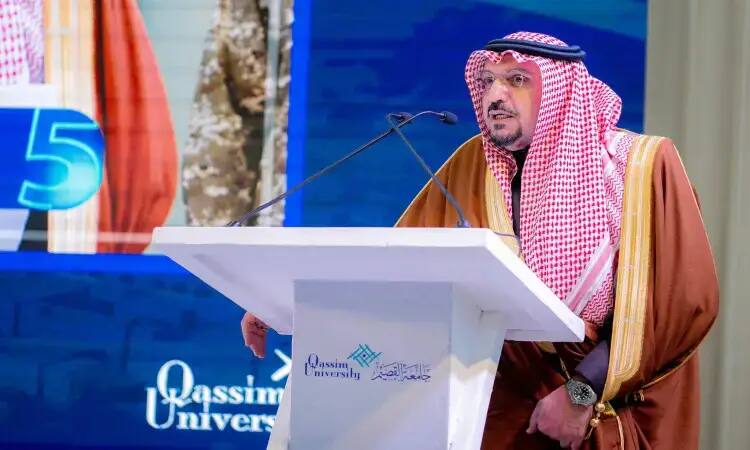The cat man, wearing the official Saudi Ghutra, Eqal and Bisht on a platform bearing the logo of Qassim University, delivers a speech in front of a prominent blue sign embodying the university's name with the number 5, highlighting the developmental balance program overseen by the Emirate of Qassim Province.