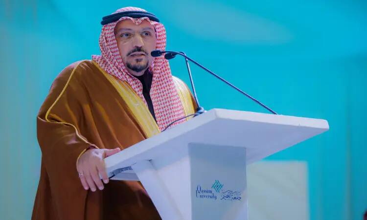 A man dressed in the Saudi Ghutra, Shamaa, Egal, and regular Saudi Bisht delivers a speech from a white podium bearing the logo of Qassim University, in front of a blue background, and the themes of awareness, health, and education, highlighting the role of Qassim Emirate in achieving developmental equilibrium.