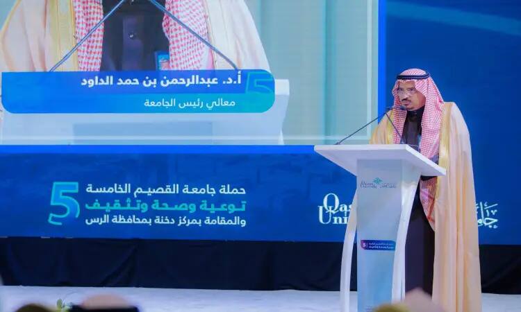 The man in the Saudi Ghutrah, Eqal and Bisht speaks from a podium with a microphone. Behind him is a large screen displaying images and text in Arabic. This comes only from Qassim University, highlighting the efforts of the Emirate of Qassim in achieving developmental vigilance, awareness and consciousness in the community.