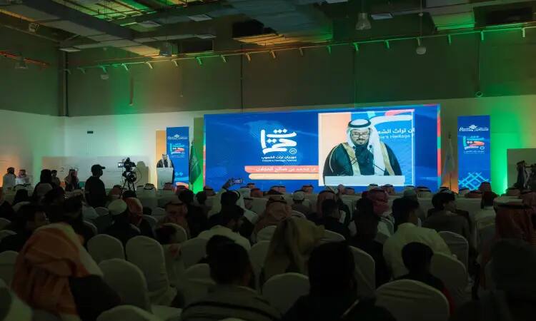 يجلس جمع غفير من الحضور في قاعة المؤتمرات، ويواجه المنصة التي يظهر عليها صاحب السمو أمير منطقة القصيم عبر الشاشة مرتديًا الغترة السعودية والعقال والبشت السعودي. سموه اختاره خلال مهرجان الجامعة، بينما تضيء أرجاء القاعة أنوار خضراء وحمراء وتلتقط صور الكاميرات لحظات الاحتفاء بتراث الشعوب.